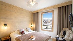 Une chambre d’hôtel avec un mur en bois, un grand lit recouvert d’une couverture à rayures, deux oreillers, un ours en peluche, une table de chevet avec un livre, une chaise, une télévision et une fenêtre donnant sur un paysage enneigé.
