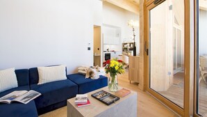 A modern living room with a blue sofa, a coffee table with books and a vase of flowers, and a view of a wooden deck through a glass door.