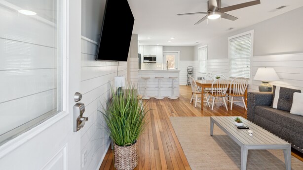 A modern living room with a flat-screen TV, a ceiling fan, a dining area with white chairs, and a grey sofa. - Swim Spa, Tiki Bar, Pet friendly, Fenced yard, 3 min walk to beach - The Blue House (Isle Of Palms)
