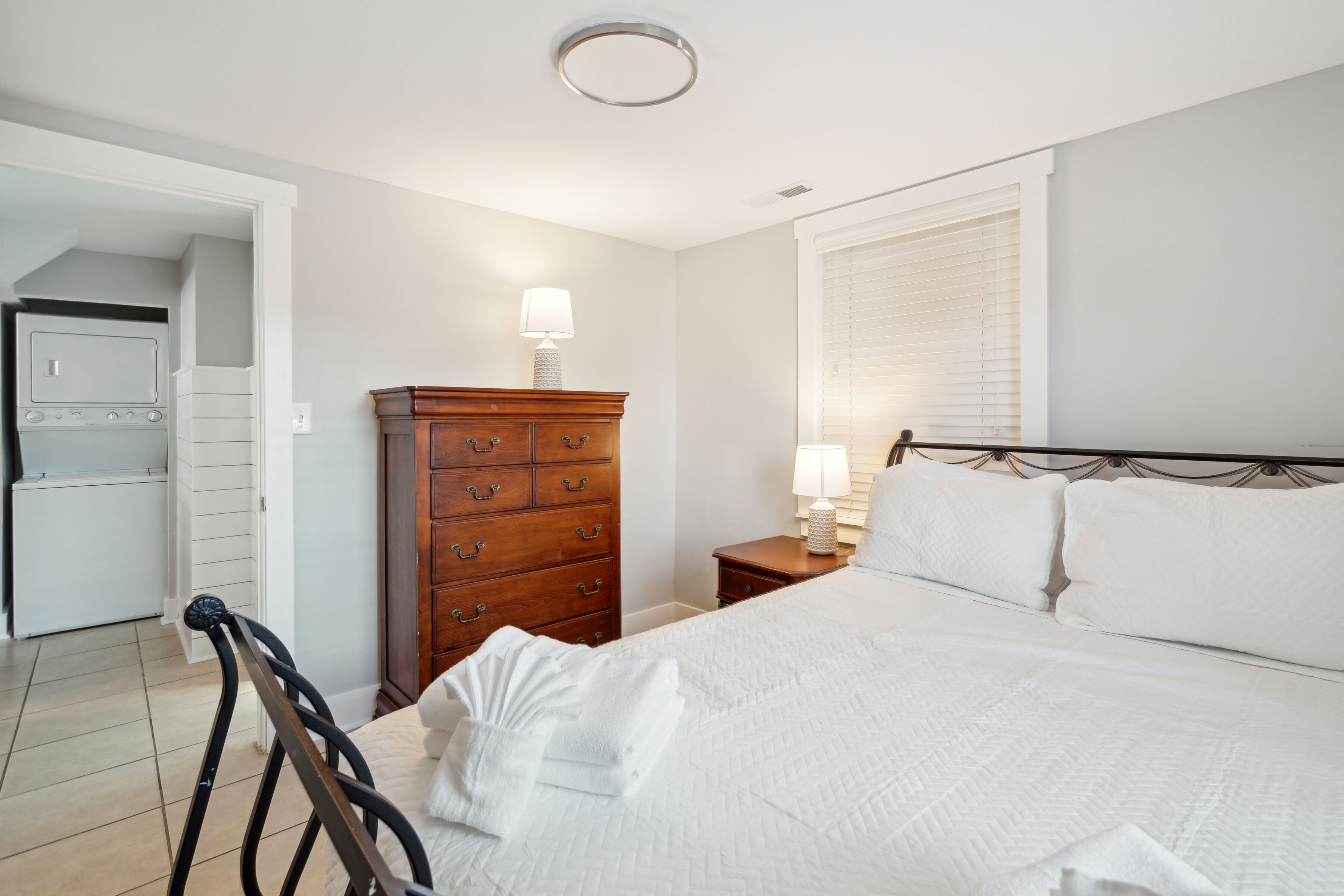 A neatly arranged bedroom with a bed, a wooden dresser, a nightstand with a lamp, and a window with blinds.