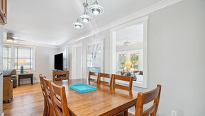 A spacious dining area with a wooden table and chairs, a television in the background, and large windows providing natural light. - Oasis w/ Private Pool, Pets considered, 2 Houses from beach access- The Pink House (Isle Of Palms)