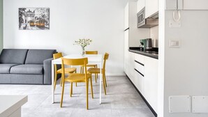 Une cuisine moderne avec des meubles blancs, une table à manger entourée de chaises jaunes et un canapé gris dans l’espace salon.