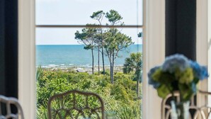 A dining area with a view of the ocean through a large window, a table with chairs, and a vase with flowers. - 7 Bedroom Oceanview Kiawah Vanderhorst Estate with Pool (Kiawah Island)