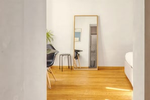 A room with a wooden floor, a bed, a chair, a small stool, a potted plant, and a mirror reflecting a staircase.
