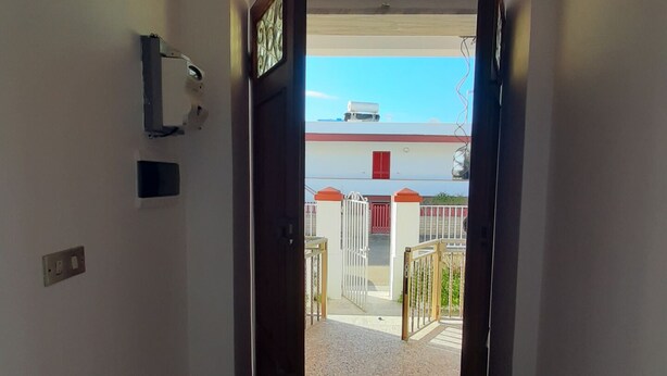 Pasillo que conduce a un área exterior amurallada, con vista despejada a un edificio con puerta roja y pared blanca.