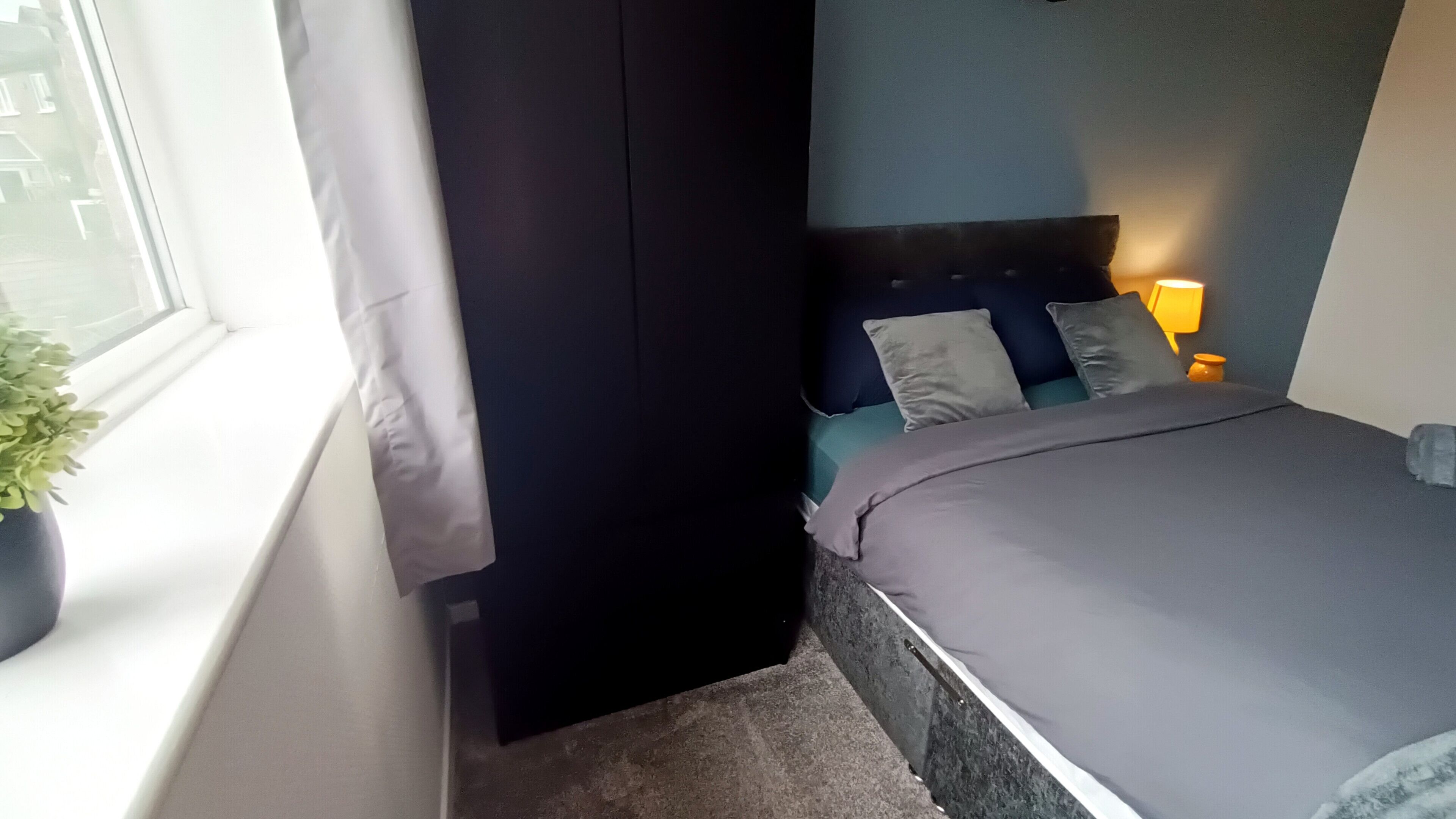 Een slaapkamer met een bed, nachtkastje met lamp, raam met gordijnen en een potplant.