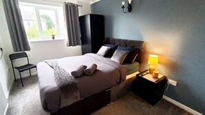 A neatly made bed with a grey blanket and two rolled towels, a black chair, a bedside table with a lamp, and a window with curtains.