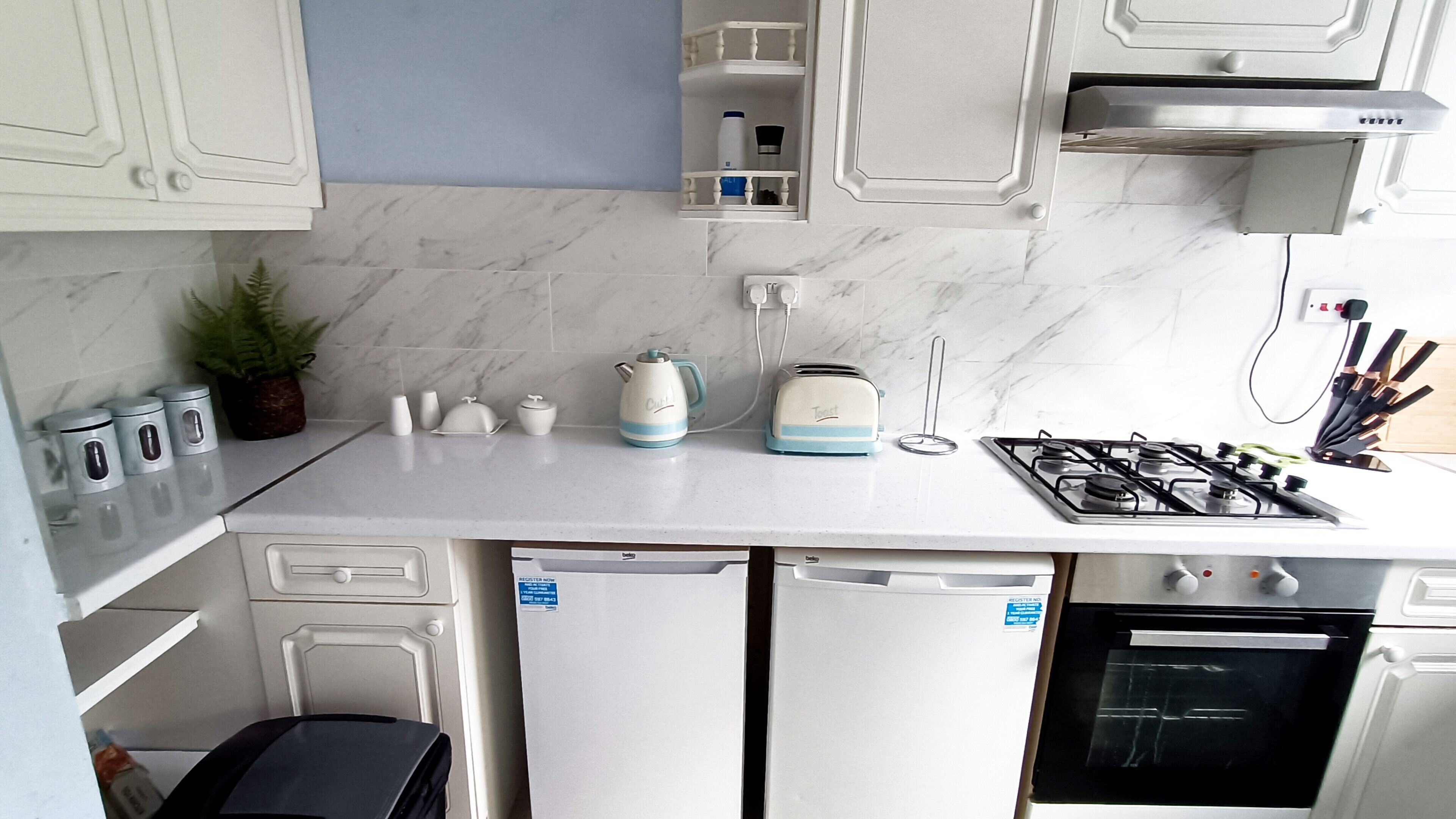 A compact kitchen with white cabinets, a marble backsplash, a gas stove, a dishwasher, and a small countertop area with kitchen appliances.