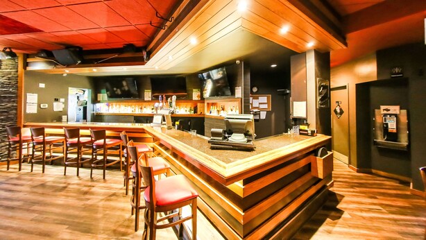 A bar area with wooden bar stools, a counter with a television, and a stone wall in the background. - Hojo House Winnipeg (Winnipeg)