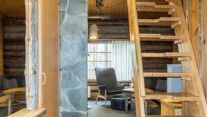 Interior rústico de una cabaña con pared de piedra, escalera de madera y un rincón acogedor con una silla y una mesa.