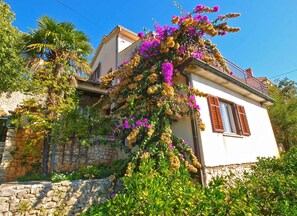 A two-story house with a stone facade, a balcony with a wooden railing, and a variety of flowering plants.