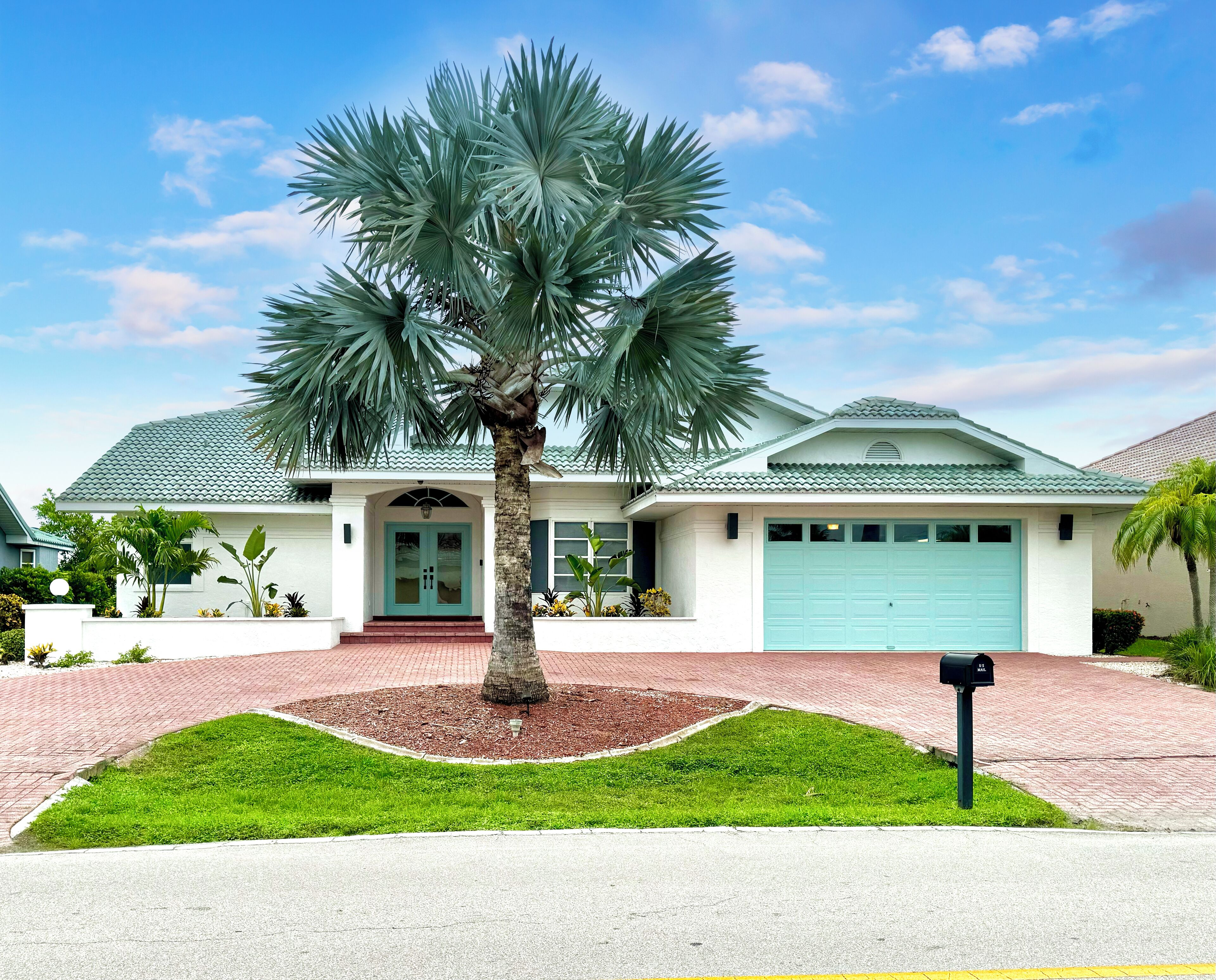 A single-story house with a light blue garage door, a central palm tree, and a brick driveway.