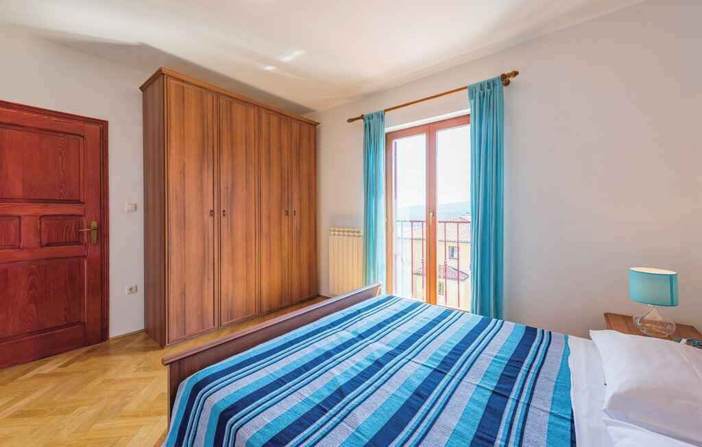 A bedroom with a bed, a wooden wardrobe, a balcony with a view, and a nightstand with a lamp.