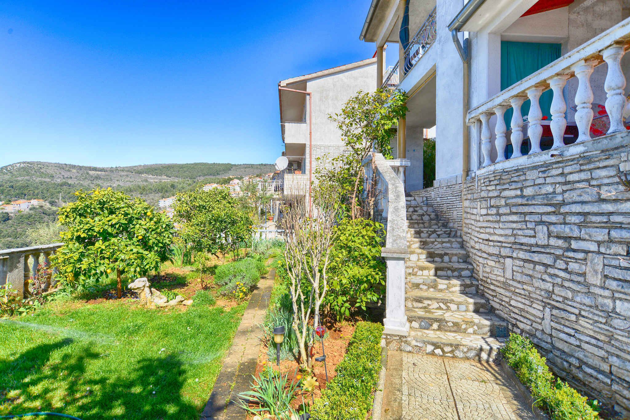 A residential building with a stone staircase, a garden with various plants, and a view of the surrounding landscape.
