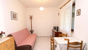 Une petite pièce bien éclairée, avec un canapé, une table entourée de chaises, un porte-manteau, un téléviseur et un meuble.