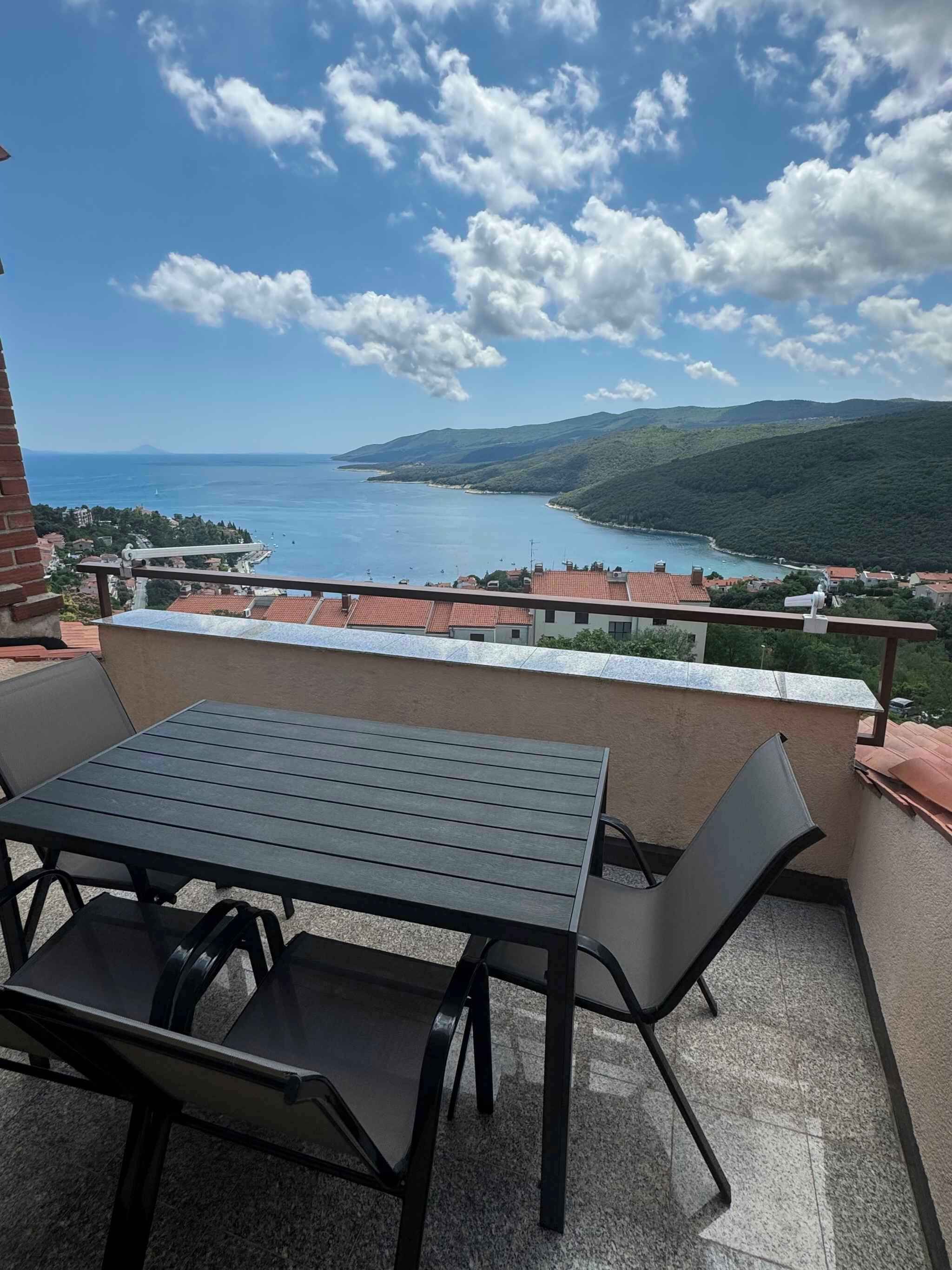 Ein Balkon mit Tisch und Stühlen mit Blick auf die Küste, Wasser und Hügel.