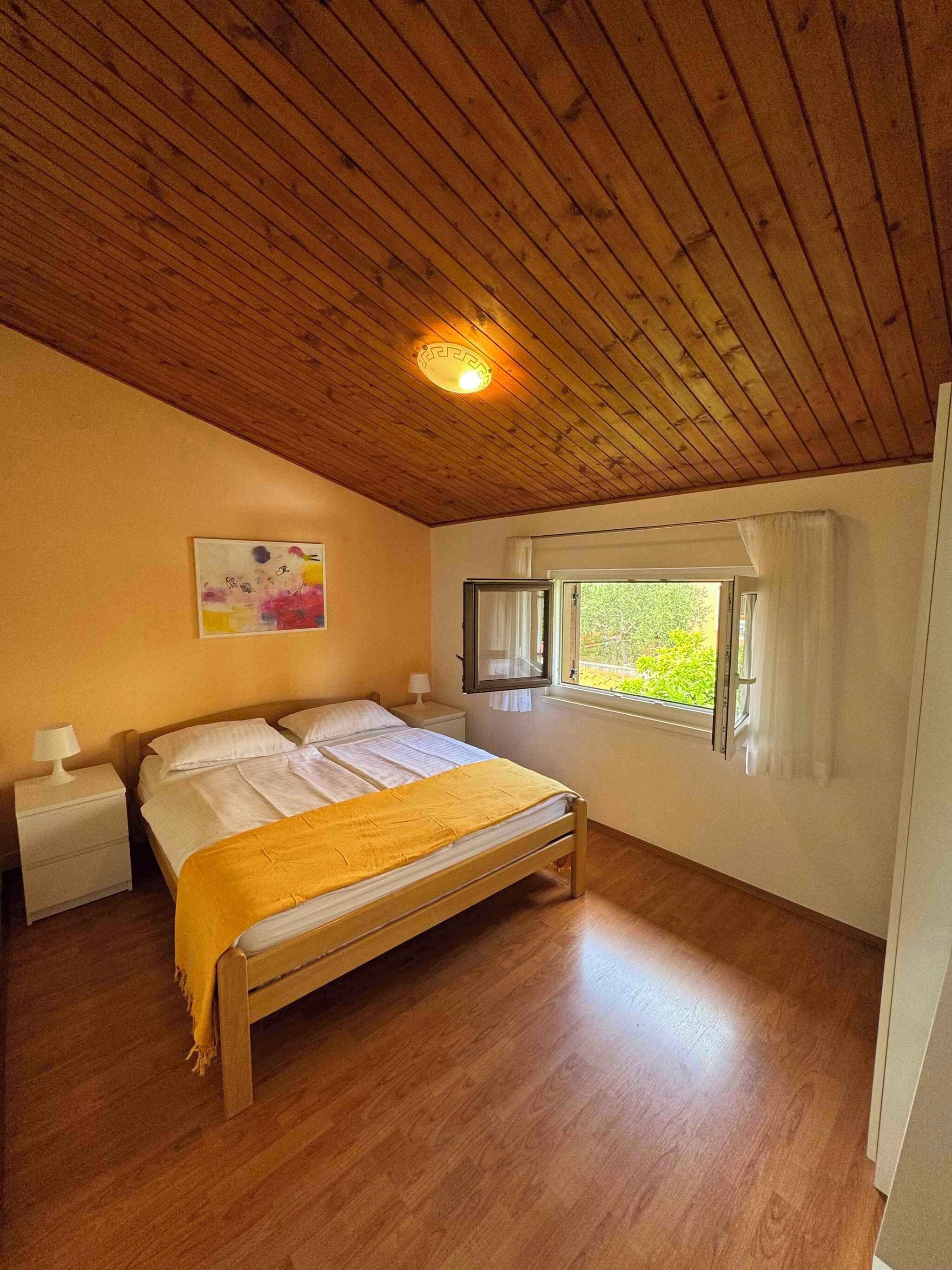 Une chambre avec un plafond en bois, un lit recouvert d’une couverture jaune, une table de chevet avec une lampe, une fenêtre avec des rideaux et un tableau accroché au mur.