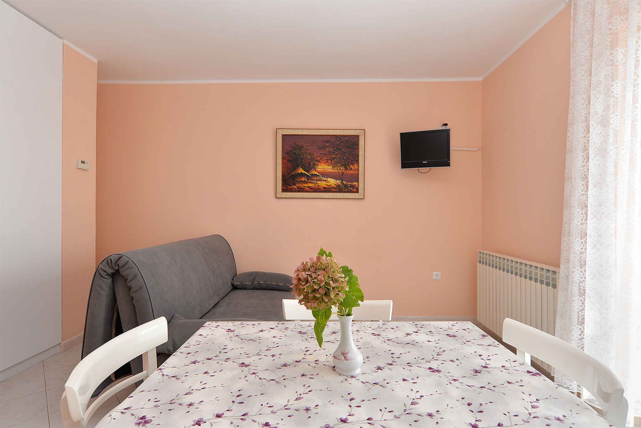 A dining area with a table set for two, a grey sofa, a painting on the wall, and a TV mounted above the sofa.