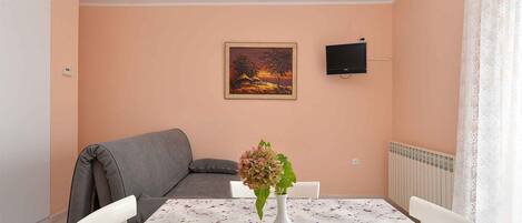 A dining area with a table set for two, a grey sofa, a painting on the wall, and a TV mounted above the sofa.