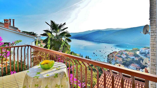 A balcony with a table, a bowl of fruit, and a view of a coastal town with boats and mountains.