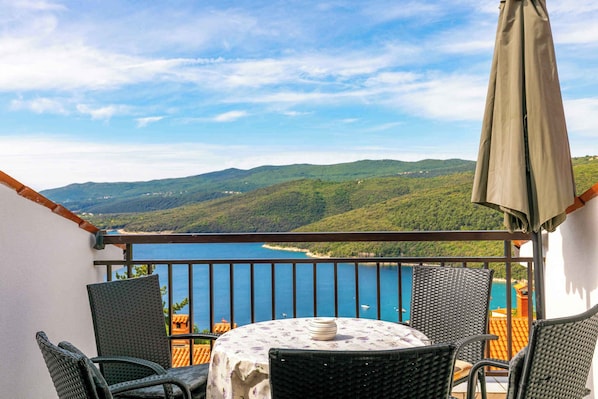 Un balcon avec des fauteuils en osier et une table, donnant sur un paysage côtier avec des collines et une étendue d’eau.