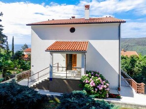 Une maison moderne à deux étages, avec un toit en tuiles, un escalier menant à l’entrée et un jardin agrémenté de fleurs roses.
