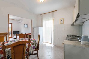 Eine Küche mit Herd, Spüle und Kühlschrank. Ein Esstisch mit Stühlen und ein Fernseher sind im Hintergrund zu sehen.