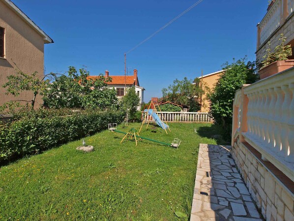 Un jardin à l’arrière d’une maison, avec une pelouse verdoyante, une balançoire et un mur en briques.