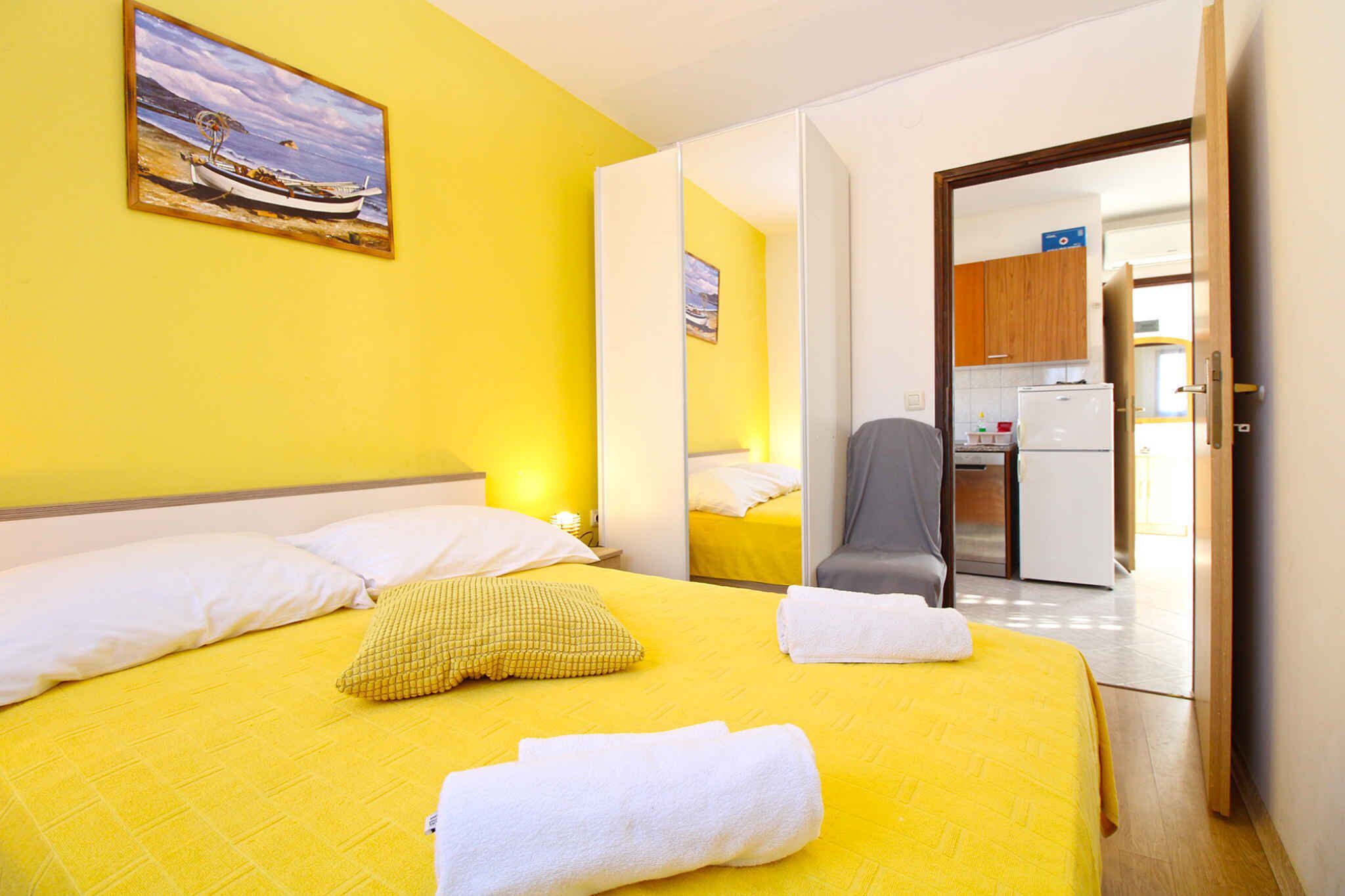 A hotel room with a yellow bedspread, white pillows, a checkered pillow, and two rolled towels. There is a painting of a boat on the wall and a view of the kitchen through an open door.