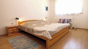 Een slaapkamer met een houten bed, een klein nachtkastje, een venster met gordijnen en een kleine stoel, plus een kleurrijk gestreept vloerkleed.
