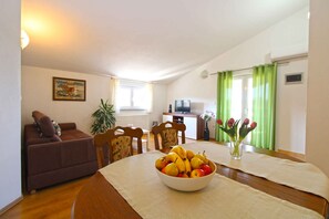 Un salon comprenant un canapé, une table à manger avec un panier de fruits, des chaises, un téléviseur et un coin cuisine.