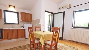A compact kitchen with wooden cabinets, a dining table with chairs, a patterned rug, and a window with blinds.