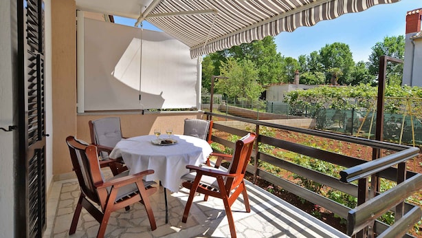 A balcony with a table set for two, a striped awning overhead, and a view of a garden.