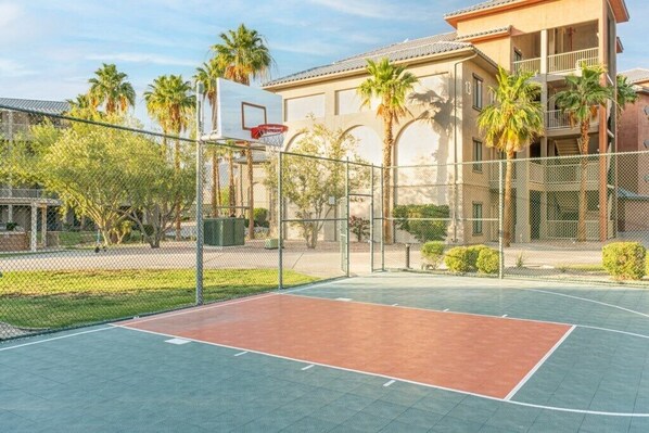 An outdoor basketball court with a basketball hoop, surrounded by a chain-link fence, palm trees, and a building with balconies.