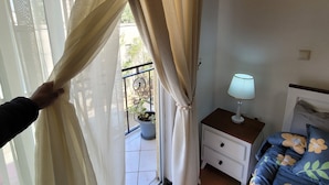 A bedroom with a bed, a nightstand, a lamp, and a view of a balcony with a potted plant. - Highly rated, clean, and spacious guest house in the Bethel area! Great discount (Kolfe Keranio)
