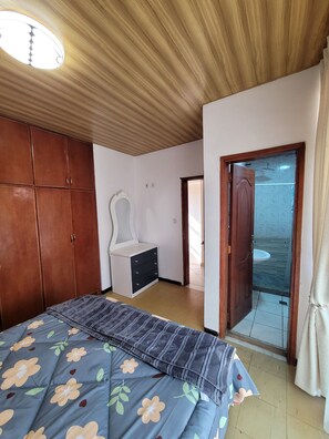 Une chambre avec une armoire en bois, un lit recouvert d’une couette à motifs floraux, une chaise blanche et une salle de bain visible à travers une porte en verre.