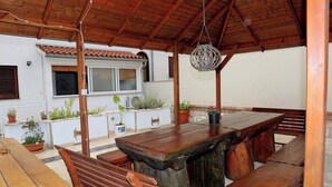 A covered outdoor dining area with wooden benches and a central table, surrounded by potted plants and a white building with a tiled roof. - Holiday apartment with air conditioning (Pula)