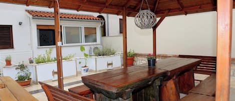 A covered outdoor dining area with wooden benches and a central table, surrounded by potted plants and a white building with a tiled roof.