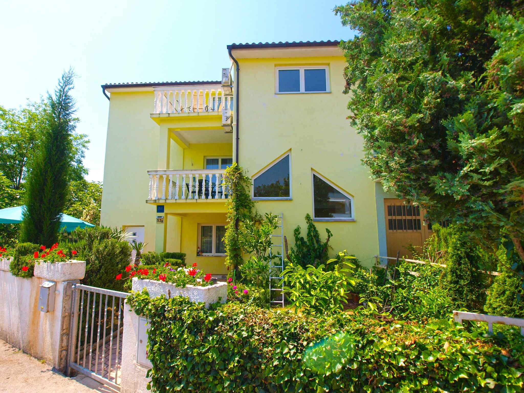 A two-story yellow building with a balcony, surrounded by lush greenery and a gated entrance.