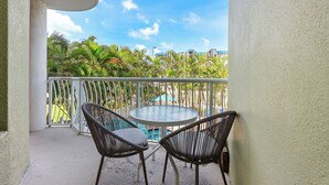 A balcony with two wicker chairs and a small round table, overlooking a pool and lush greenery.