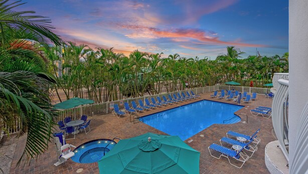 A pool area with blue lounge chairs and umbrellas, surrounded by palm trees and a view of the sky.