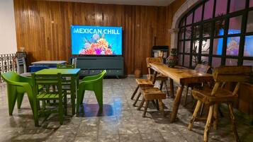 A room with a wooden wall, a television displaying 'MEXICAN CHILL,' a table with chairs, and a window with a view.