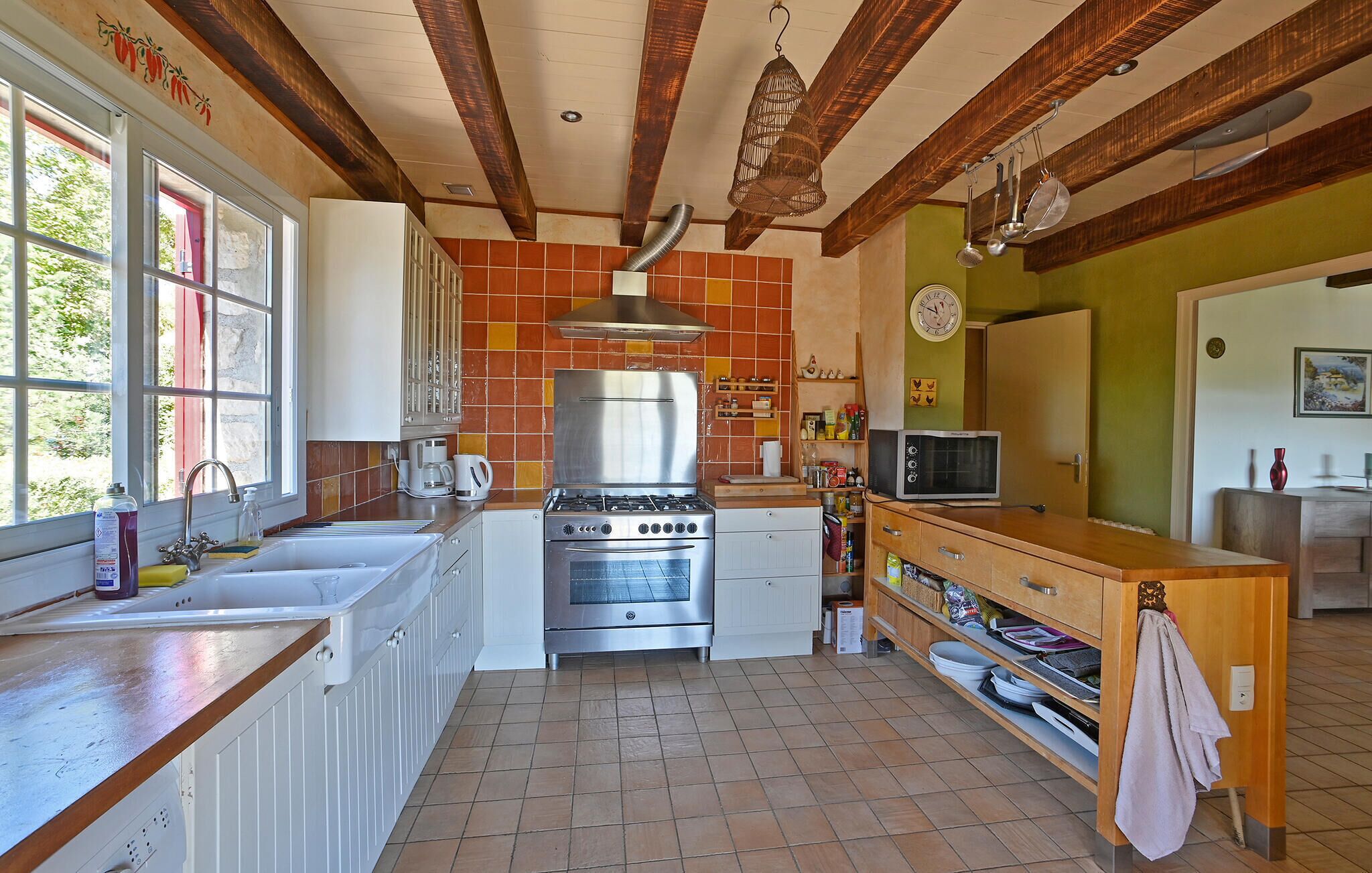 A kitchen with a stove, oven, and microwave, wooden beams on the ceiling, and a window with a view of greenery.