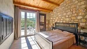 Een slaapkamer met een stenen muur, een houten plafond, een bed, een nachtkastje en uitzicht door een glazen deur op een buitenruimte.