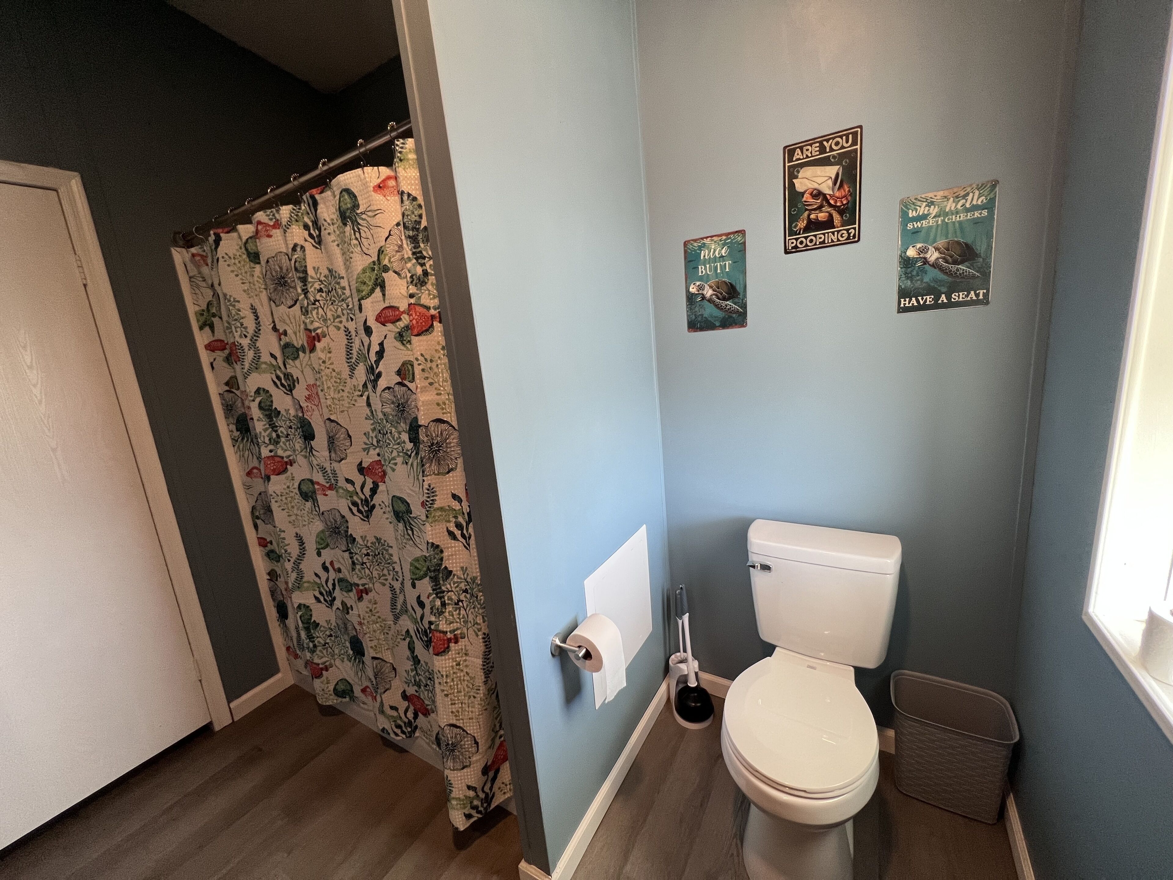 A bathroom with a toilet, a shower curtain with a floral pattern, and three wall-mounted signs with humorous messages.