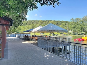 A riverside outdoor seating area with tables and chairs, a covered pavilion, and a fenced walkway along the water.