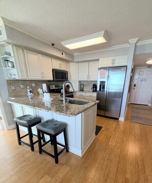 A kitchen with white cabinets, a granite countertop, stainless steel appliances, and two bar stools.