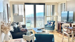 A living room with a wooden floor, a sofa, a blue armchair, a coffee table, a TV, and a view through large glass doors. - Two of the three bedrooms have ocean front balconies! (North Myrtle Beach)