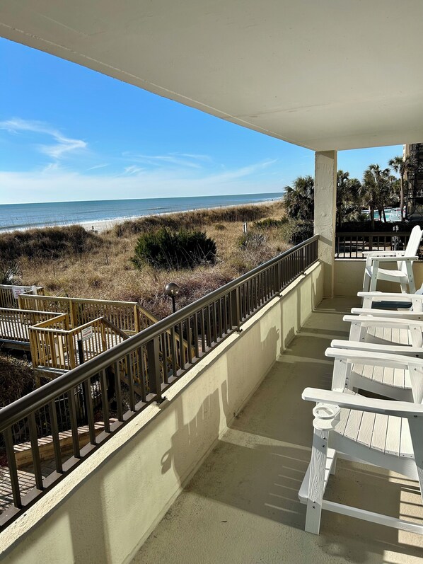 A balcony with white Adirondack chairs, a wooden railing, and a view of the beach and ocean.