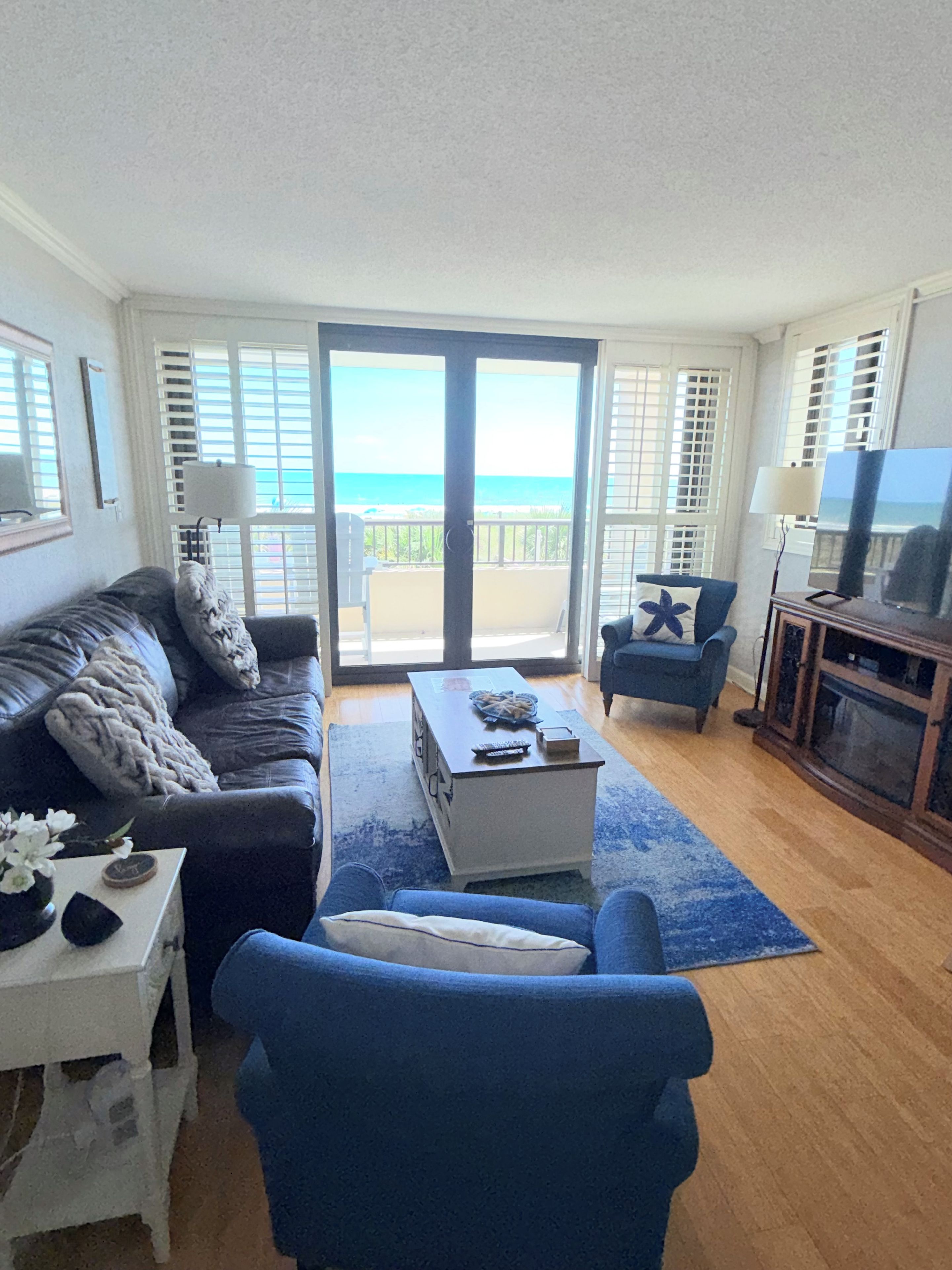 A living room with a sofa, a coffee table, and a TV. There is a balcony with a view of the ocean.